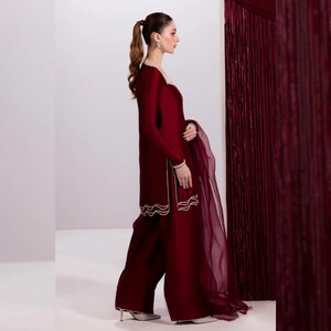 May include: A burgundy-colored ensemble featuring a long-sleeved tunic and wide-leg pants. The tunic has white embroidered trim along the neckline, cuffs, and hem. A sheer burgundy scarf drapes from the arm. The outfit is paired with silver heels.