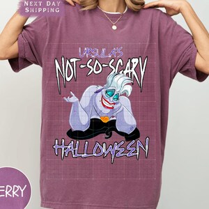 May include: Berry-colored t-shirt featuring a graphic of Ursula from The Little Mermaid. The design includes the text "Ursula's Not-So-Scary Halloween" in a spooky font, with Ursula's image in the center.