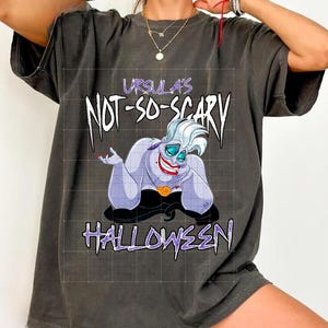 May include: Dark gray t-shirt featuring a cartoon image of Ursula from The Little Mermaid. The text "Ursula's Not-So-Scary Halloween" is printed in white and purple. The shirt is a relaxed fit.