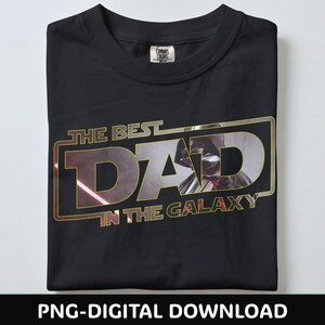 Może przedstawiać: Czarny t-shirt ze złotą i srebrną grafiką z napisem "THE BEST DAD IN THE GALAXY". Grafika przedstawia sylwetkę Dartha Vadera. Koszulka jest starannie złożona na białej powierzchni.