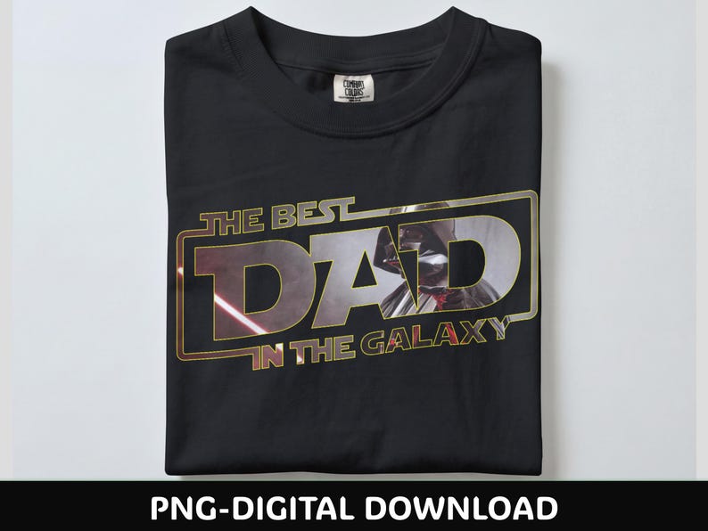 Może przedstawiać: Czarny t-shirt ze złotą i srebrną grafiką z napisem "THE BEST DAD IN THE GALAXY". Grafika przedstawia sylwetkę Dartha Vadera. Koszulka jest starannie złożona na białej powierzchni.