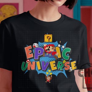 May include: Black t-shirt featuring a colorful graphic design with the words "EPIC UNIVERSE" in a comic book style. The design includes pixelated characters and elements from a popular video game.
