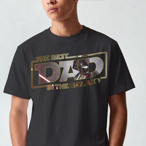 Może przedstawiać: Czarny t-shirt z napisem "THE BEST DAD IN THE GALAXY" w kolorze złotym i czerwonym. Projekt zawiera wizerunek postaci z popularnej serii science fiction. Koszulka ma klasyczny okrągły dekolt.