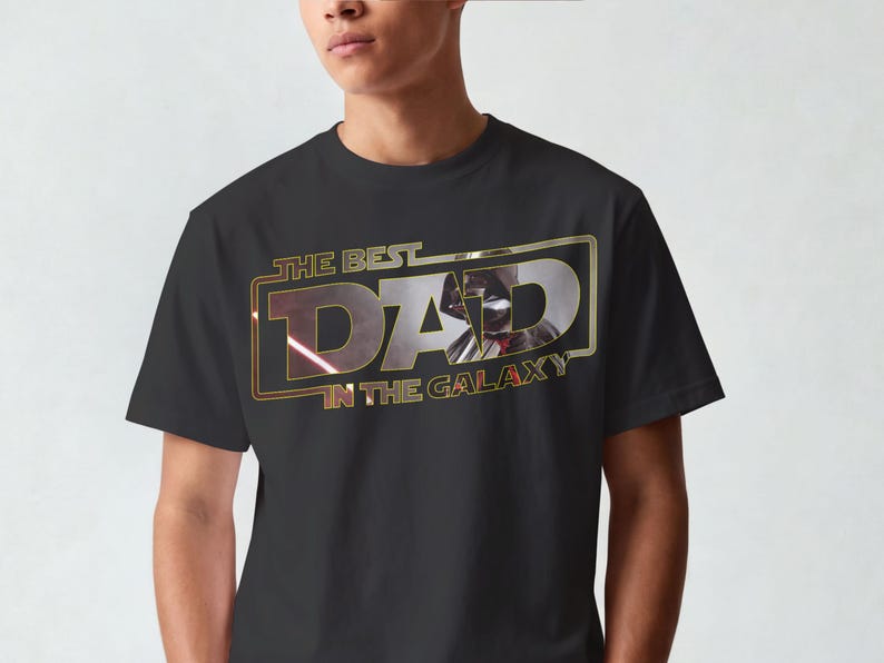 Może przedstawiać: Czarny t-shirt z napisem "THE BEST DAD IN THE GALAXY" w kolorze złotym i czerwonym. Projekt zawiera wizerunek postaci z popularnej serii science fiction. Koszulka ma klasyczny okrągły dekolt.