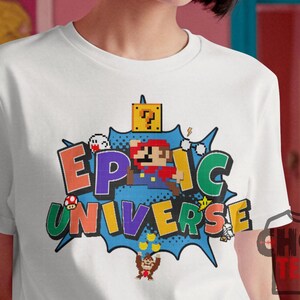 May include: White t-shirt featuring a colorful graphic design with the words "EPIC UNIVERSE" in pixelated letters. The design includes pixelated characters and elements from a popular video game.