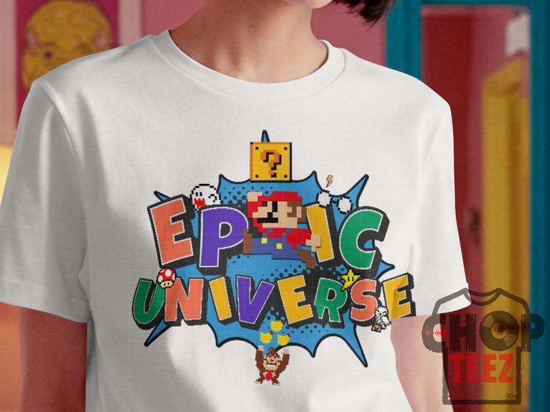 May include: White t-shirt featuring a colorful graphic design with the words "EPIC UNIVERSE" in pixelated letters. The design includes pixelated characters and elements from a popular video game.