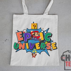 May include: White tote bag featuring a colorful graphic of a pixelated Mario character and the words "EPIC UNIVERSE" in a comic book style. The design includes various game elements and characters.