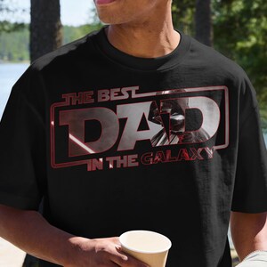 Może przedstawiać: Czarny t-shirt z napisem "THE BEST DAD IN THE GALAXY" w kolorze czerwonym i białym. Grafika przedstawia sylwetkę Dartha Vadera. Koszulka ma okrągły dekolt i wydaje się być wykonana z bawełny.