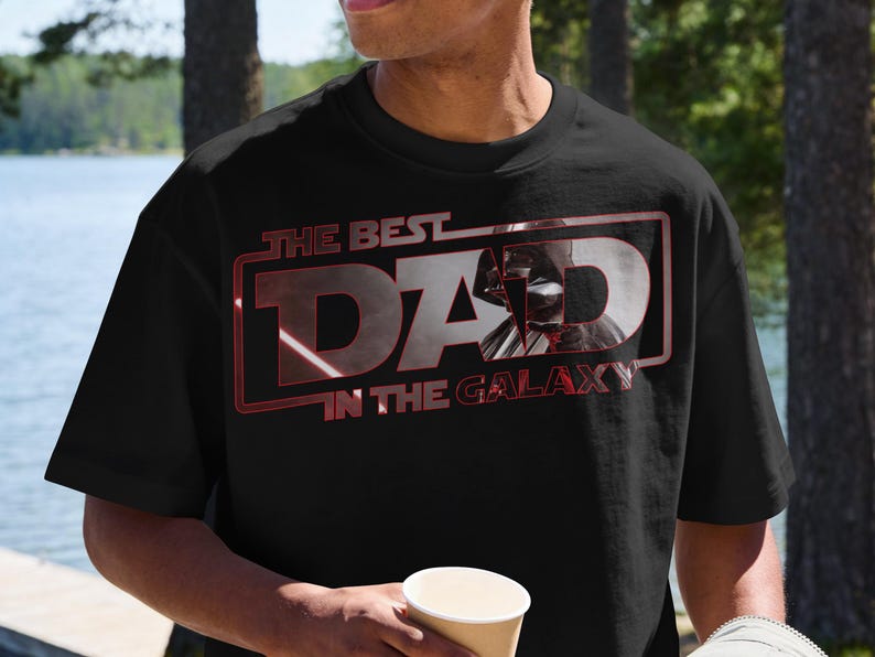 Może przedstawiać: Czarny t-shirt z napisem "THE BEST DAD IN THE GALAXY" w kolorze czerwonym i białym. Grafika przedstawia sylwetkę Dartha Vadera. Koszulka ma okrągły dekolt i wydaje się być wykonana z bawełny.