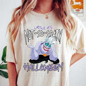 May include: Cream-colored t-shirt featuring a cartoon illustration of Ursula from The Little Mermaid. The graphic includes the text "Ursula's Not-So-Scary Halloween" in purple and black. The character has purple skin, a red smile, and a black dress.