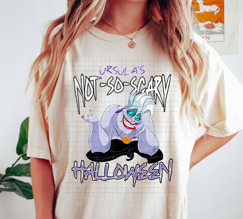 May include: Cream-colored t-shirt featuring a cartoon illustration of Ursula from The Little Mermaid. The graphic includes the text "Ursula's Not-So-Scary Halloween" in purple and black. The character has purple skin, a red smile, and a black dress.