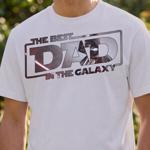 Może przedstawiać: Biała koszulka z napisem "THE BEST DAD IN THE GALAXY" w prostokątnym wzorze. Wz&oacute;r przedstawia metaliczną srebrno-czerwoną grafikę hełmu Dartha Vadera. Koszulka jest z kr&oacute;tkim rękawem.