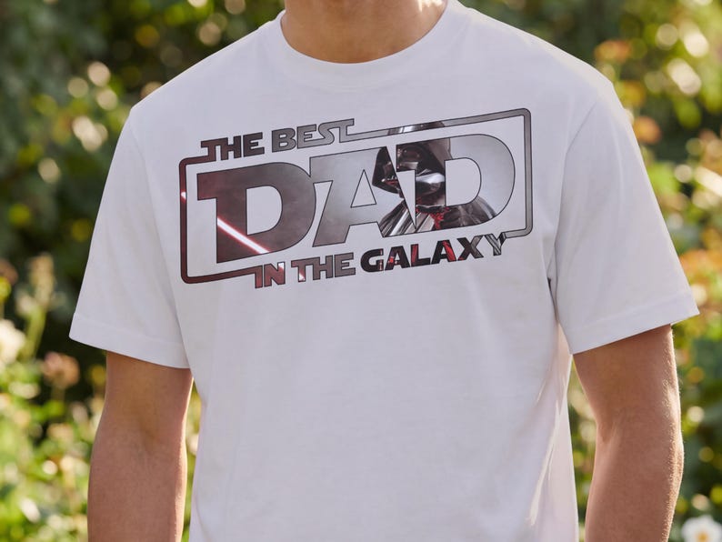 Może przedstawiać: Biała koszulka z napisem "THE BEST DAD IN THE GALAXY" w prostokątnym wzorze. Wz&oacute;r przedstawia metaliczną srebrno-czerwoną grafikę hełmu Dartha Vadera. Koszulka jest z kr&oacute;tkim rękawem.
