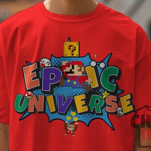 May include: Red t-shirt featuring a pixelated graphic with the words "EPIC UNIVERSE" in colorful letters. The design includes pixelated characters and elements from a popular video game, set against a blue burst background.