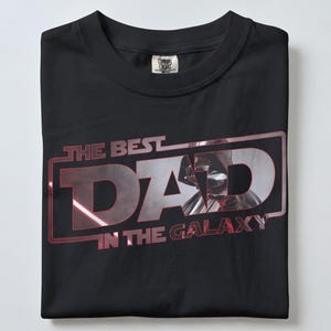 Może przedstawiać: Czarny t-shirt z napisem "THE BEST DAD IN THE GALAXY" w kolorze czerwonym i srebrnym. Grafika przedstawia stylizowany wizerunek Dartha Vadera. Koszulka jest starannie złożona.