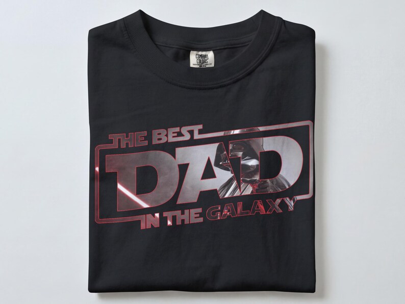 Może przedstawiać: Czarny t-shirt z napisem "THE BEST DAD IN THE GALAXY" w kolorze czerwonym i srebrnym. Grafika przedstawia stylizowany wizerunek Dartha Vadera. Koszulka jest starannie złożona.
