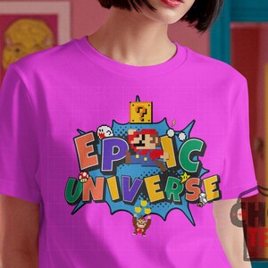 May include: A magenta t-shirt featuring a colorful graphic design. The design includes the words "EPIC UNIVERSE" in a comic book style, along with pixelated characters and elements from a popular video game.