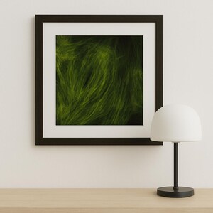Op de afbeelding: Een ingelijste abstracte kunstafdruk met een donkerbruine lijst, met een levendig groen, getextureerd ontwerp. Een wit-koepelvormige tafellamp met een zwarte voet en steel staat op een lichtgekleurd houten oppervlak.
