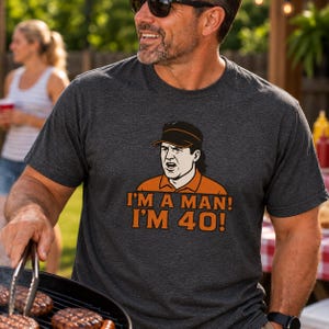 May include: A charcoal gray short-sleeve t-shirt featuring a graphic illustration of a person wearing a visor and an orange polo shirt. Below the illustration, the text reads I'M A MAN! I'M 40! in bold orange capital letters on the front of the shirt.