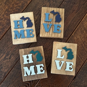 以下が含まれることがあります： 「HOME」と「LOVE」の文字が様々な青と白で書かれた4つの木製ブロック。各ブロックにはミシガン州の切り抜きが施されています。ブロックは濃い木材の表面に配置されています。