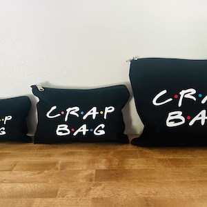 May include: Three black zippered pouches of different sizes, each with the white text "C.R.A.P. BAG" in a stylized font with coloured dots. They are arranged on a wooden surface.