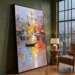 May include: An abstract oil painting in a black frame depicts a waterscape with boats and buildings. The artwork features a vibrant palette of colors, including yellow, orange, blue, and gray, creating a textured, impressionistic effect. The painting is leaning against a green wall.