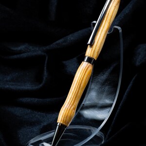 May include: A ballpoint pen featuring a light brown wood grain barrel with a smooth, contoured shape. The pen includes polished dark metal components, including a pointed tip, a center band, and a pocket clip attached to the cap.