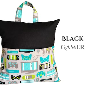 Puede incluir: Una almohada de lectura negra con asa, con un estampado de mandos de videojuegos grises, turquesas y negros en la mitad inferior. La mitad superior es negra lisa, con el texto "BLACK GAMER" a la derecha.