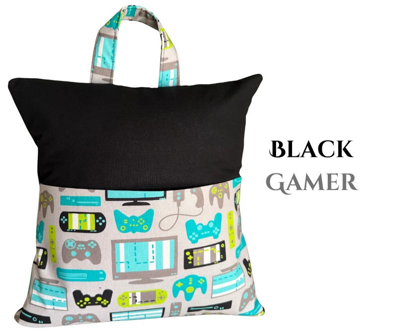 Puede incluir: Una almohada de lectura negra con asa, con un estampado de mandos de videojuegos grises, turquesas y negros en la mitad inferior. La mitad superior es negra lisa, con el texto "BLACK GAMER" a la derecha.