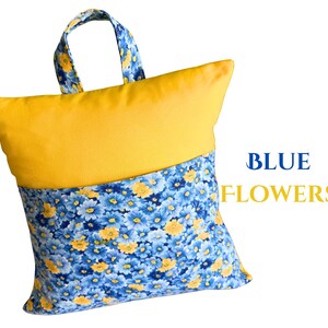 Puede incluir: Un coj&iacute;n amarillo y azul con asa. La parte superior es amarilla lisa, mientras que la inferior presenta un estampado floral azul con detalles amarillos. El texto "BLUE FLOWERS" se muestra en azul y amarillo.