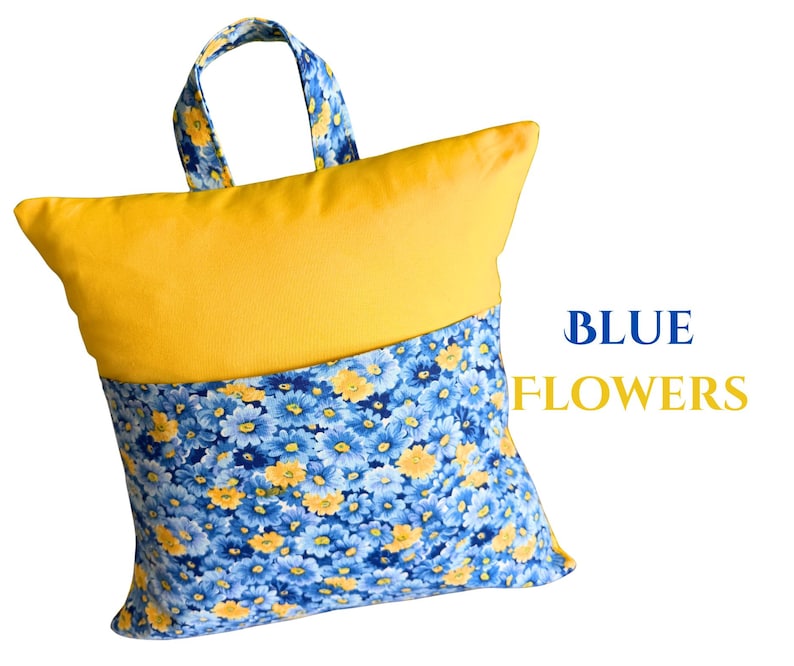 Puede incluir: Un coj&iacute;n amarillo y azul con asa. La parte superior es amarilla lisa, mientras que la inferior presenta un estampado floral azul con detalles amarillos. El texto "BLUE FLOWERS" se muestra en azul y amarillo.