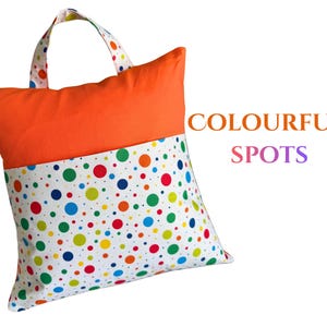 Puede incluir: Un coj&iacute;n cuadrado con asa, con una secci&oacute;n superior naranja vibrante y una secci&oacute;n inferior blanca adornada con lunares de colores. El texto "COLOURFUL SPOTS" se muestra en el lado derecho del coj&iacute;n.