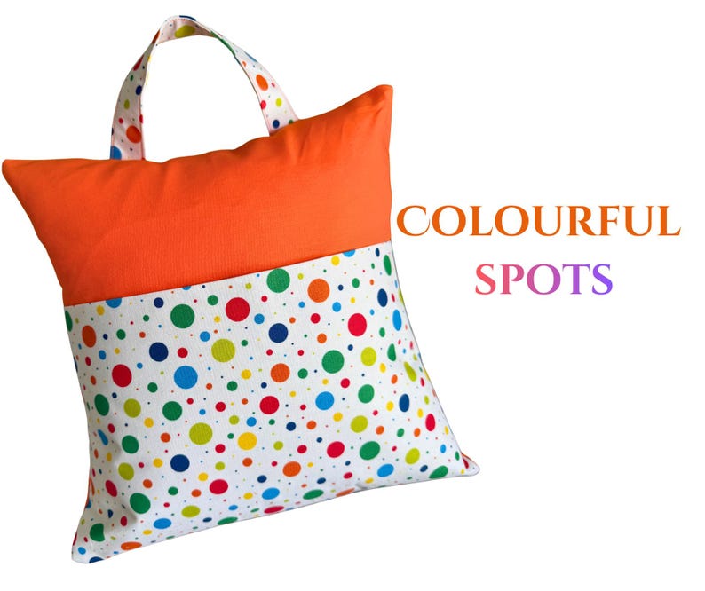 Puede incluir: Un coj&iacute;n cuadrado con asa, con una secci&oacute;n superior naranja vibrante y una secci&oacute;n inferior blanca adornada con lunares de colores. El texto "COLOURFUL SPOTS" se muestra en el lado derecho del coj&iacute;n.