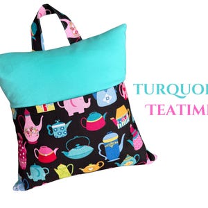 Puede incluir: Una almohada de lectura turquesa con una tela base negra estampada con coloridas teteras. La almohada tiene una parte superior turquesa y un asa con un estampado de tetera. El texto "TURQUOISE TEATIME" es visible.