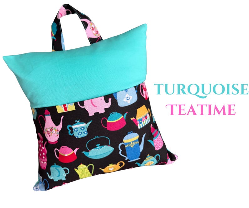 Puede incluir: Una almohada de lectura turquesa con una tela base negra estampada con coloridas teteras. La almohada tiene una parte superior turquesa y un asa con un estampado de tetera. El texto "TURQUOISE TEATIME" es visible.