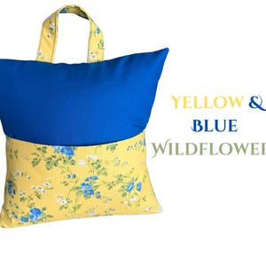Puede incluir: Una almohada de lectura con la parte superior azul y la inferior floral amarilla. El asa y la parte inferior de la almohada presentan un estampado floral amarillo con flores azules y blancas. El texto "YELLOW & BLUE WILDFLOWERS" est&aacute; a la derecha.