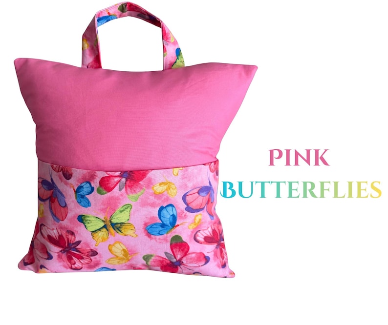 Puede incluir: Una almohada de lectura rosa con asa. La parte superior es rosa liso, mientras que la inferior presenta un estampado de mariposas rosas con mariposas rojas, azules y amarillas. El texto "PINK BUTTERFLIES" se muestra en la imagen.