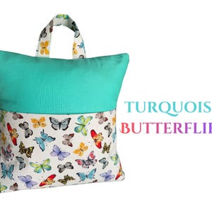 Puede incluir: Una almohada de lectura turquesa y blanca con asa. La parte superior es turquesa lisa, mientras que la inferior presenta una tela blanca estampada con mariposas de colores. El texto "TURQUOISE BUTTERFLIES" es visible a la derecha.