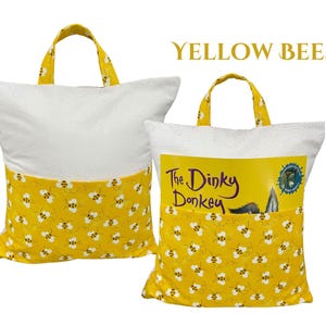 Puede incluir: Dos almohadas para libros blancas y amarillas con asas. La parte inferior de las almohadas y las asas son amarillas con un estampado de abejas. Una almohada tiene un libro en el bolsillo que dice "The Dinky Donkey". El texto "YELLOW BEES" est&aacute; en la parte superior.