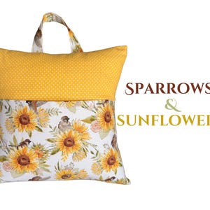 Puede incluir: Una almohada de lectura amarilla con asa. La secci&oacute;n superior es amarilla con lunares blancos. La secci&oacute;n inferior presenta un estampado floral de girasoles y gorriones sobre un fondo blanco. El texto "SPARROWS & SUNFLOWERS" est&aacute; a la derecha.