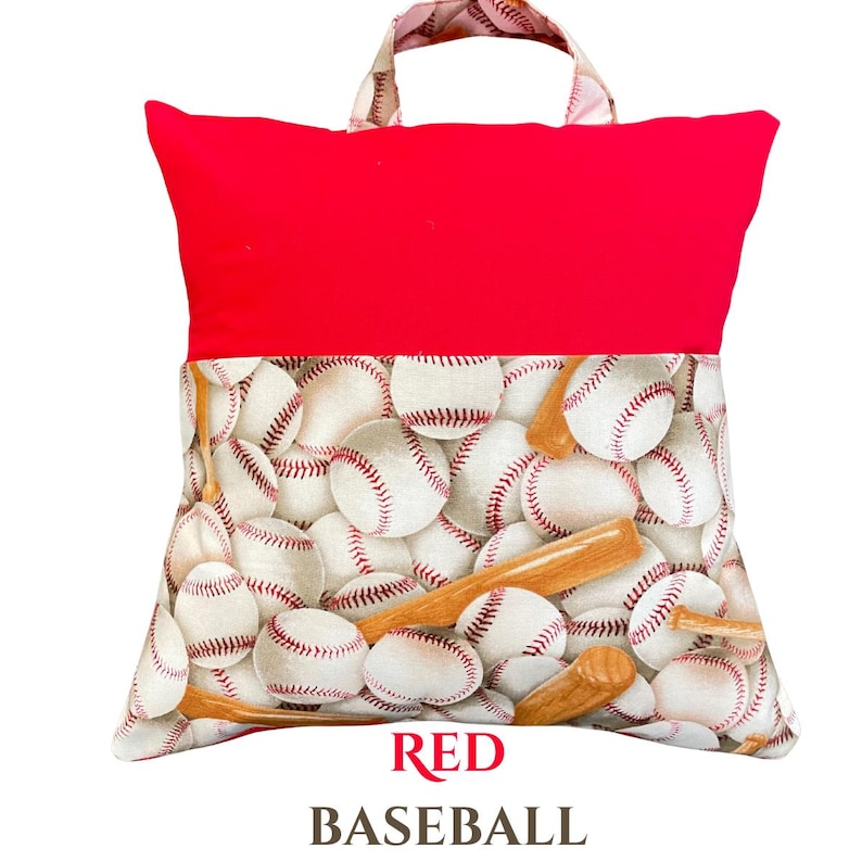 Puede incluir: Una almohada de lectura roja con asa. La secci&oacute;n superior es de color rojo liso, mientras que la inferior presenta un estampado de pelotas de b&eacute;isbol blancas y bates de madera. El texto "RED BASEBALL" est&aacute; impreso en la parte inferior.