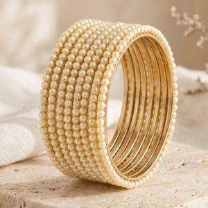 May include: A set of seven stacked gold-toned metal bangles, each featuring a single row of small, round, cream-colored faux pearls embedded along the outer surface, creating a uniform and textured appearance across the entire stack.