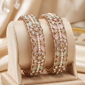 May include: Two gold-tone metal bangles featuring a central row of small white pearls flanked by alternating marquise-shaped light pink and mint green stones, with clear round crystals set in a decorative filigree pattern throughout the bands.