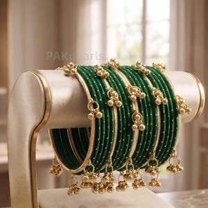 May include: A set of dark green bangles featuring a smooth texture with small gold-colored dots and dangling gold-colored bell charms. The bangles are displayed on a cylindrical stand, and the text PAKpearls is visible in the background.