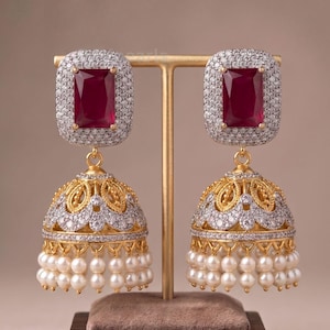 May include: A pair of gold-tone jhumka earrings featuring a rectangular deep red stone at the top, surrounded by clear pavé crystals. The bell-shaped base is decorated with intricate gold filigree, clear crystals, and a row of small white pearls.