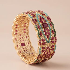 May include: A wide gold-tone bangle bracelet featuring an intricate filigree pattern. The surface is embellished with marquise-cut red stones, small round turquoise beads, and tiny green accents arranged in a repeating floral-inspired design.