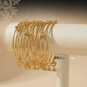 May include: A collection of gold-colored bangles displayed on a white stand. Each bangle features small, dangling gold-colored beads. The bangles are arranged in a curved line, showcasing their design and craftsmanship. The background is a soft, neutral tone.