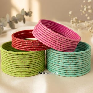 May include: A stack of colorful bangles in shades of pink, red, green, and teal. The bangles have a metallic finish with small gold-colored dots. The bangles are stacked in a neat arrangement, with a soft, neutral background.