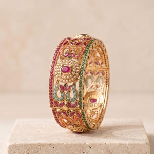 May include: A gold-colored bangle bracelet with intricate floral and geometric designs. The bracelet is adorned with red, turquoise, and green gemstones. The bracelet is displayed on a light-colored surface.