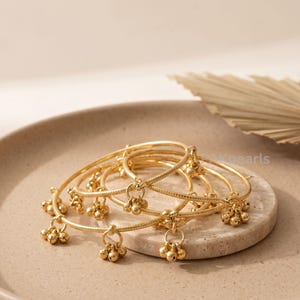 May include: A collection of gold-colored bangles with small bell charms. The bangles are arranged on a neutral-toned surface, with a few bangles in the foreground and others stacked in the background. The bells are clustered in groups of three.